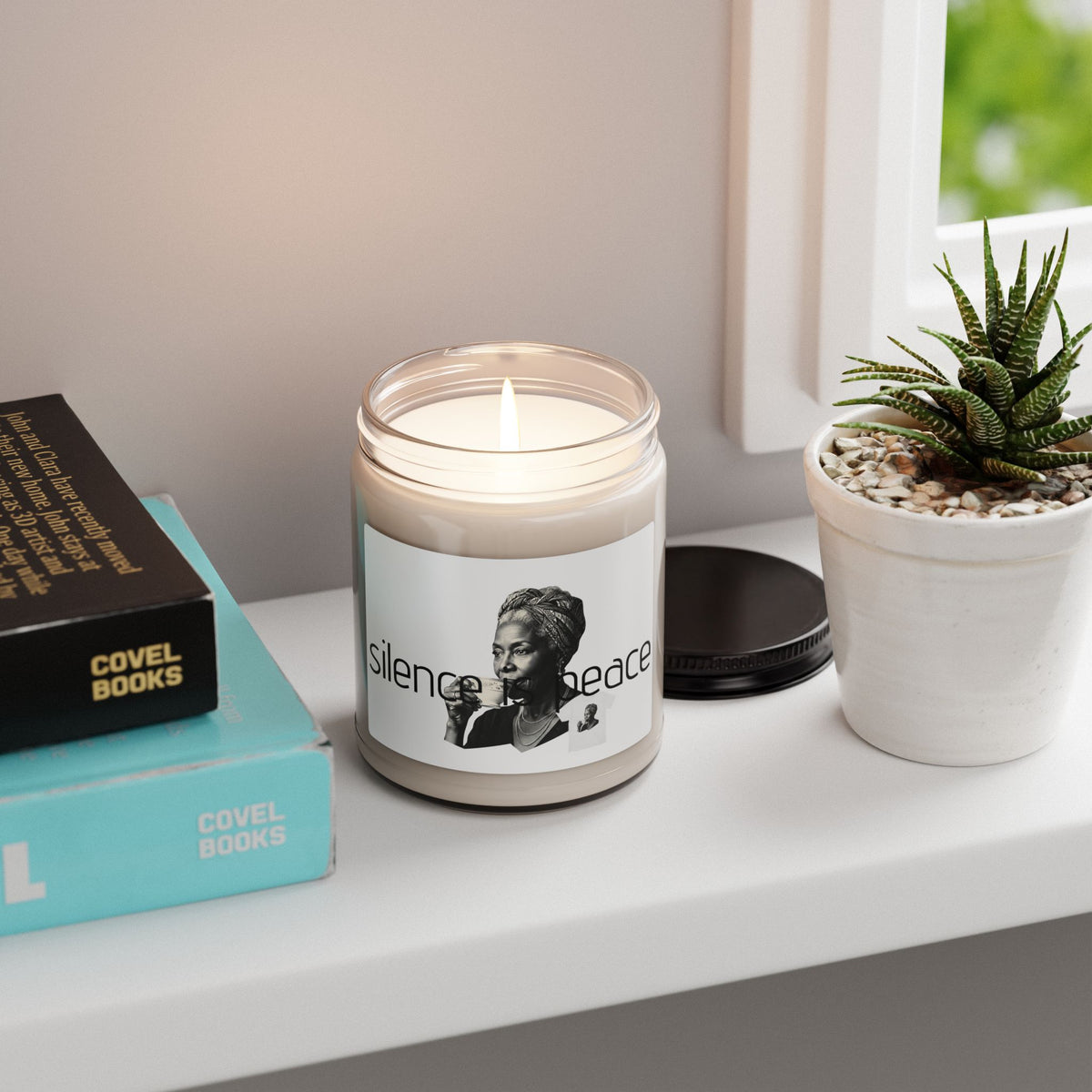 A lit candle in a clear glass jar sits on a white windowsill next to a small potted succulent and stacked books The candle label reads, "silence peace with black- white portraits illustration 