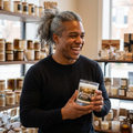 a smiling person with a gray hair in a ponytail holds a candle in a shop with decor blurred in the bckground 