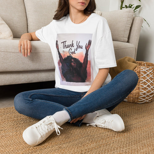 A woman sitting cross-legged on a woven rug in front of a beige sofa, wearing a white graphic T-shirt with the text 'Thank You God' and a dark portrait print, blue jeans, and white sneakers.