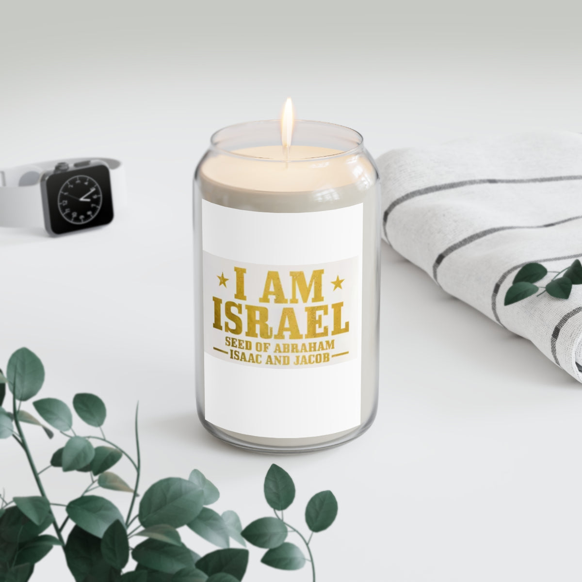 A glass jar candle with a white label reading "I AM ISRAEL" in gold lettering styled on a light surface with a folded towel and with a watch in the background