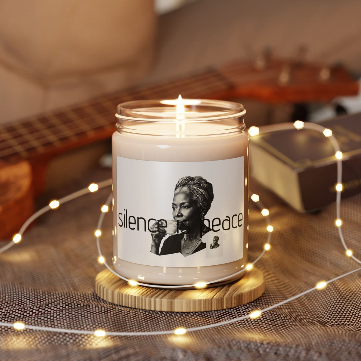 A lit  in a clear glass jar sits on a wooden coaster, surrounded by warm white fairy lights featuring a black-and-white portraits of a woman and the words "silence peace" in black text